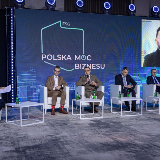 zdjęcie kolorowe. Na krzesłąch postawionych na scenie siedzi pięć osób obok siebie. Obok siedzi osoba, która prowadzi dyskusję